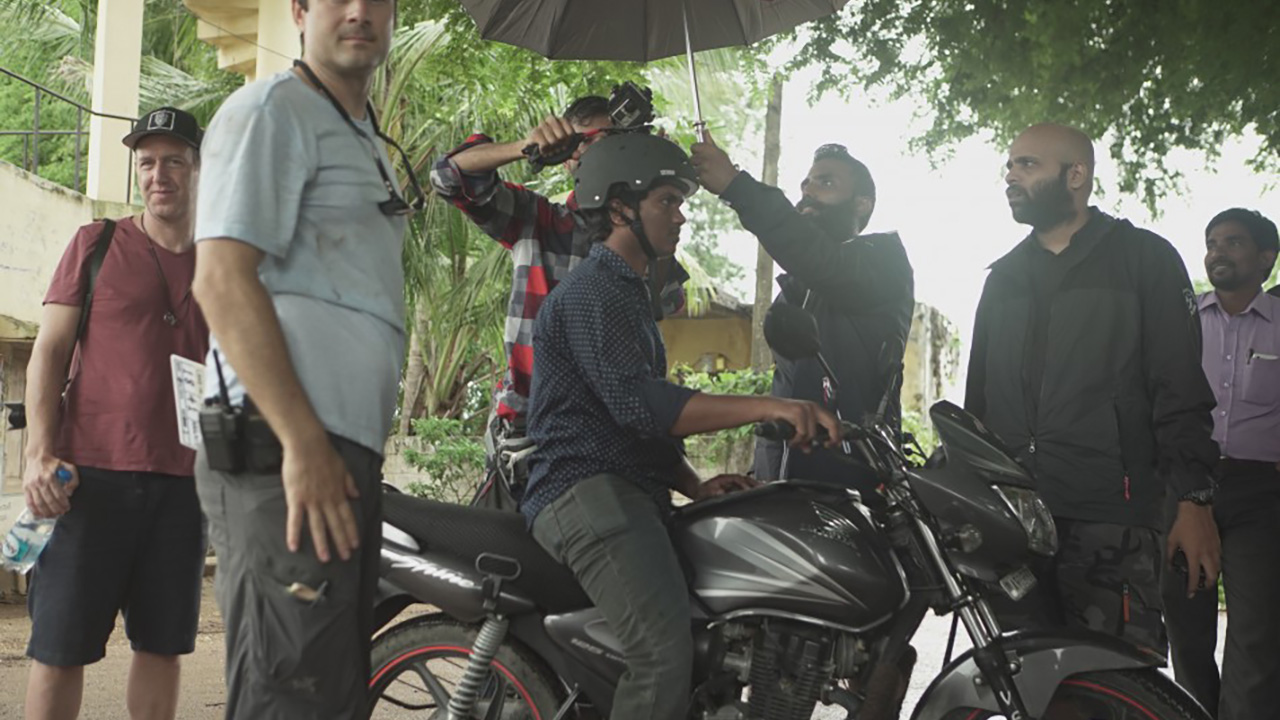 THE RETURN: Crew film a helmeted man on a motorbike, holding an umbrella over the camera rig on an overcast day.