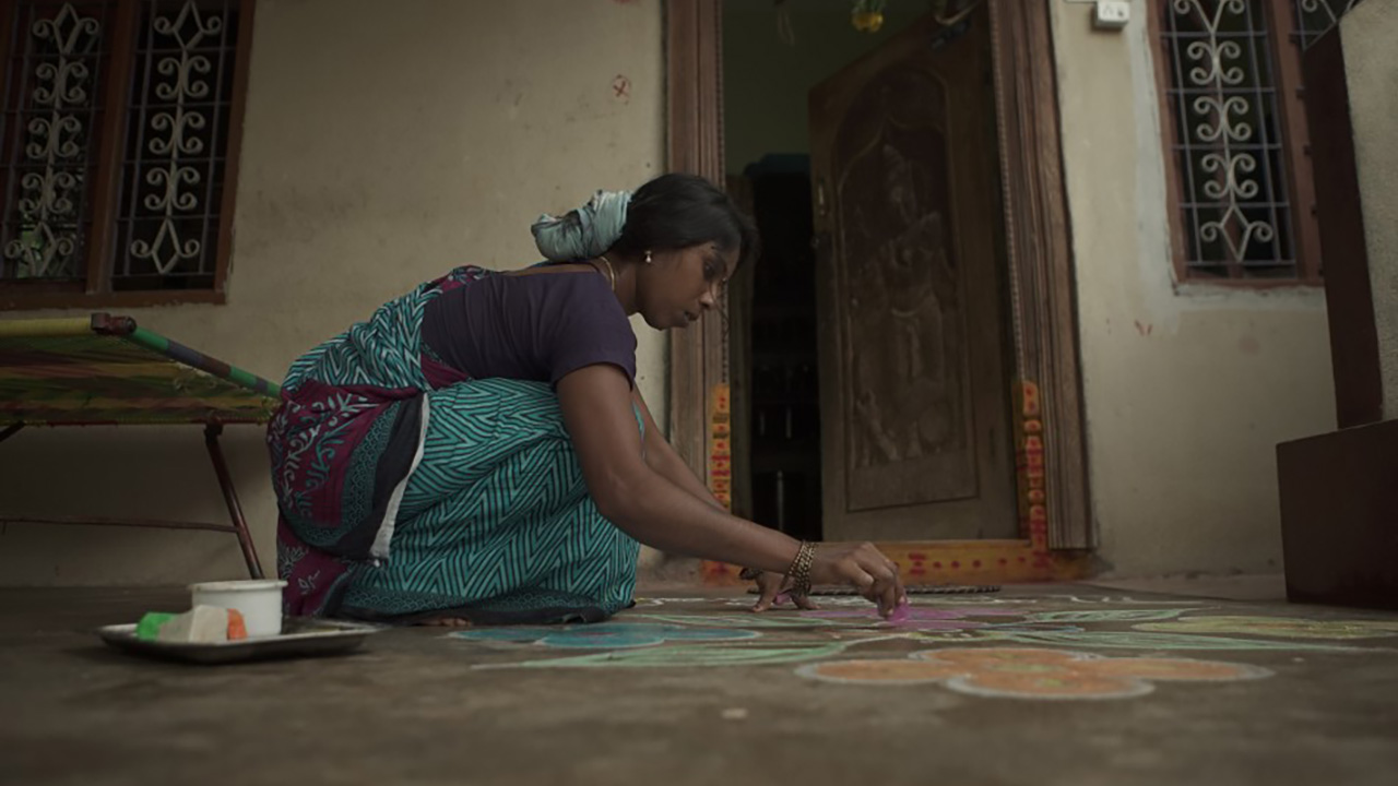 THE RETURN: A woman kneels to draw a colourful rangoli pattern on the ground in front of a doorway.