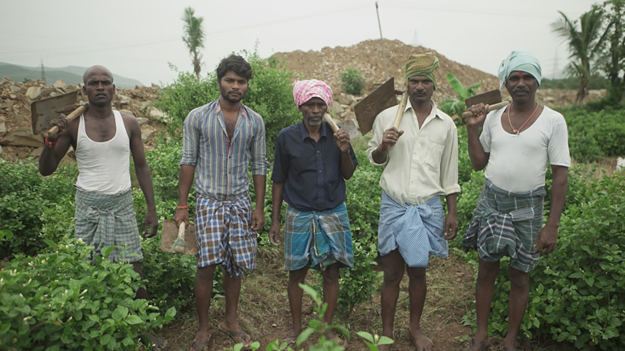 THE RETURN: Five men stand in a green field, each holding a mattock over their shoulder.