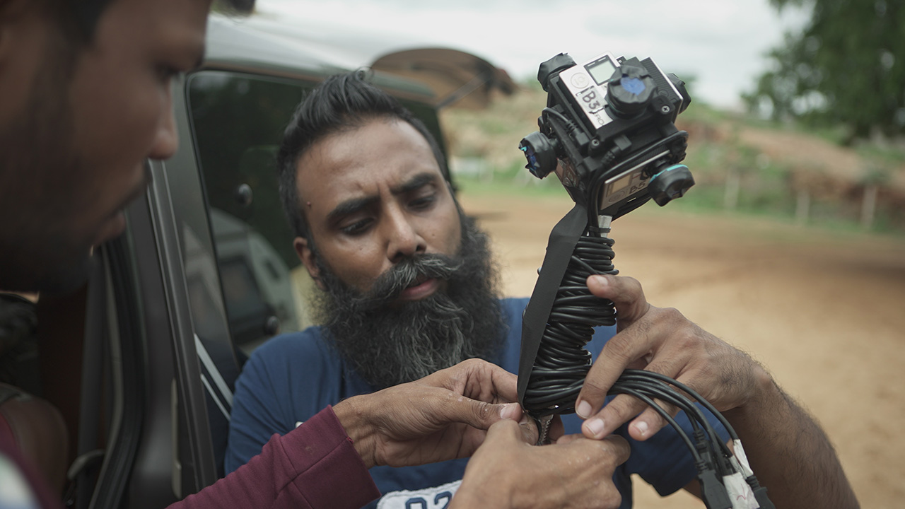 THE RETURN: Two crew members rig a 360° camera with trailing cables beside a car.