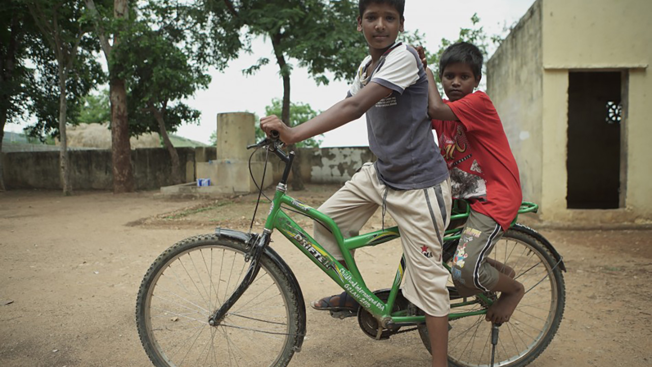 THE RETURN: Two boys ride together on a green bicycle along a dirt path.