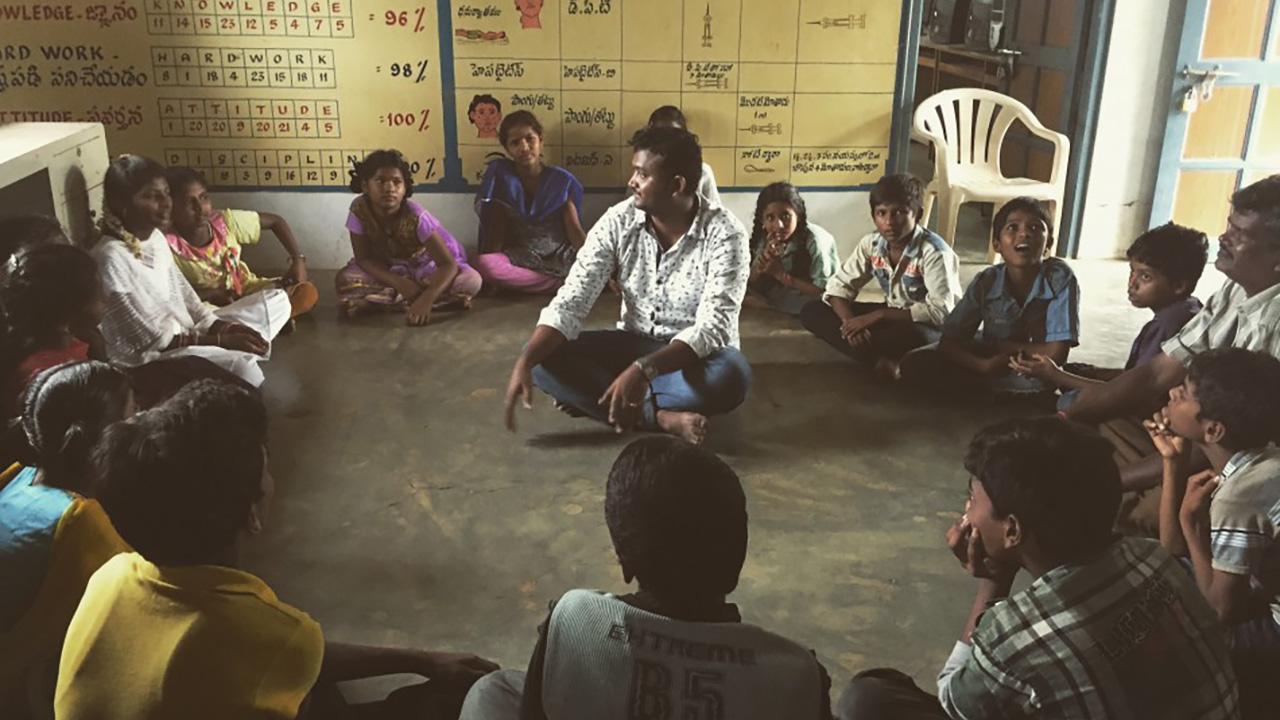 The Return: a man sitting cross legged at the center of a circle of children and teenagers, speaking to them in a community room decorated with educational charts on knowledge, hard work, attitude and discipline.