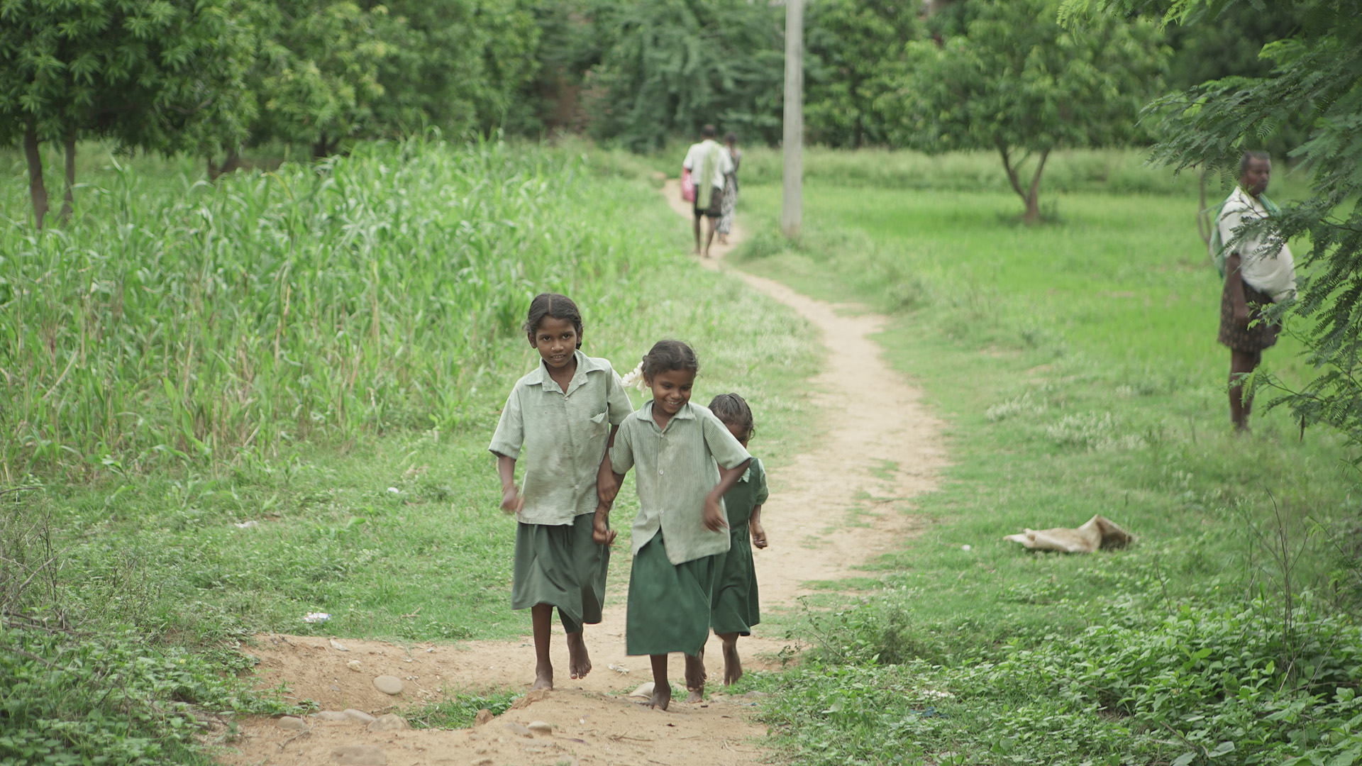 The Return: three girls in green and white school uniforms walking hand in hand along a dirt path through green fields, with adults and a resting dog visible along the path.