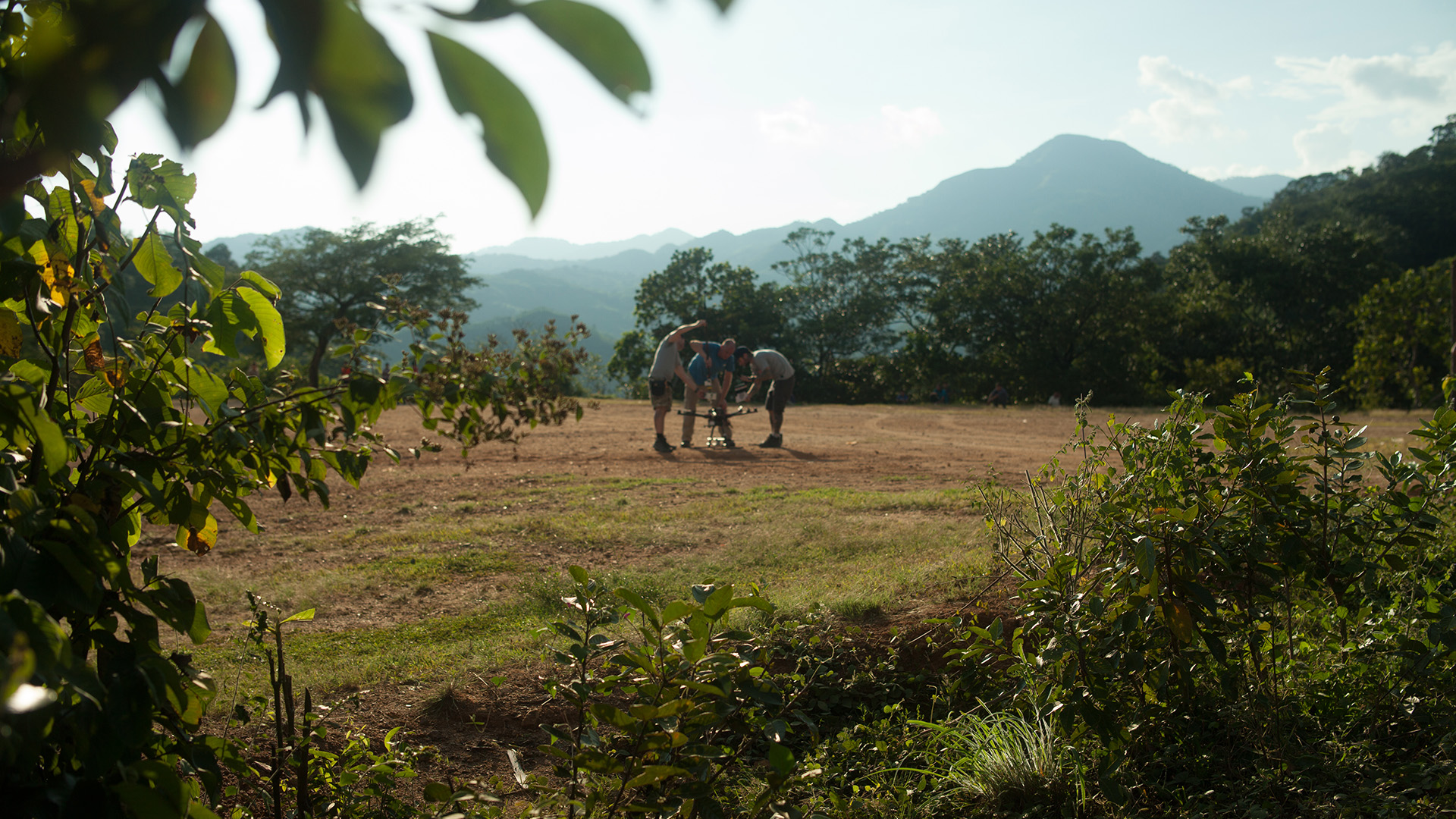 WATER: Three crew members set up a drone in a sunlit field in rural Honduras, mountains rising in the background.