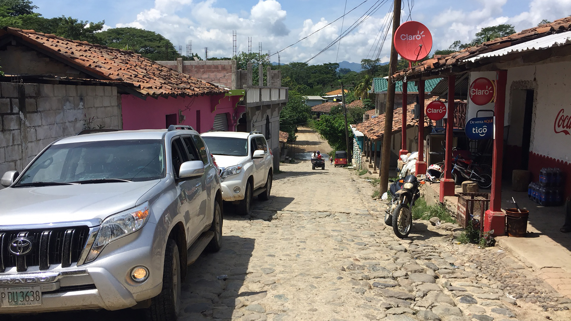 WATER: A cobblestone street in a Honduran village lined with tile-roofed houses, parked SUVs, and a motorbike.